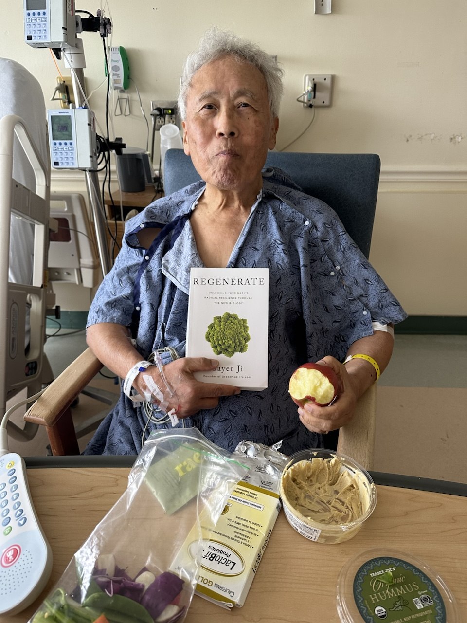 Dr. Ji in the ICU holding his son Sayer Ji's book Regenerate, with fresh vegetables and hummus